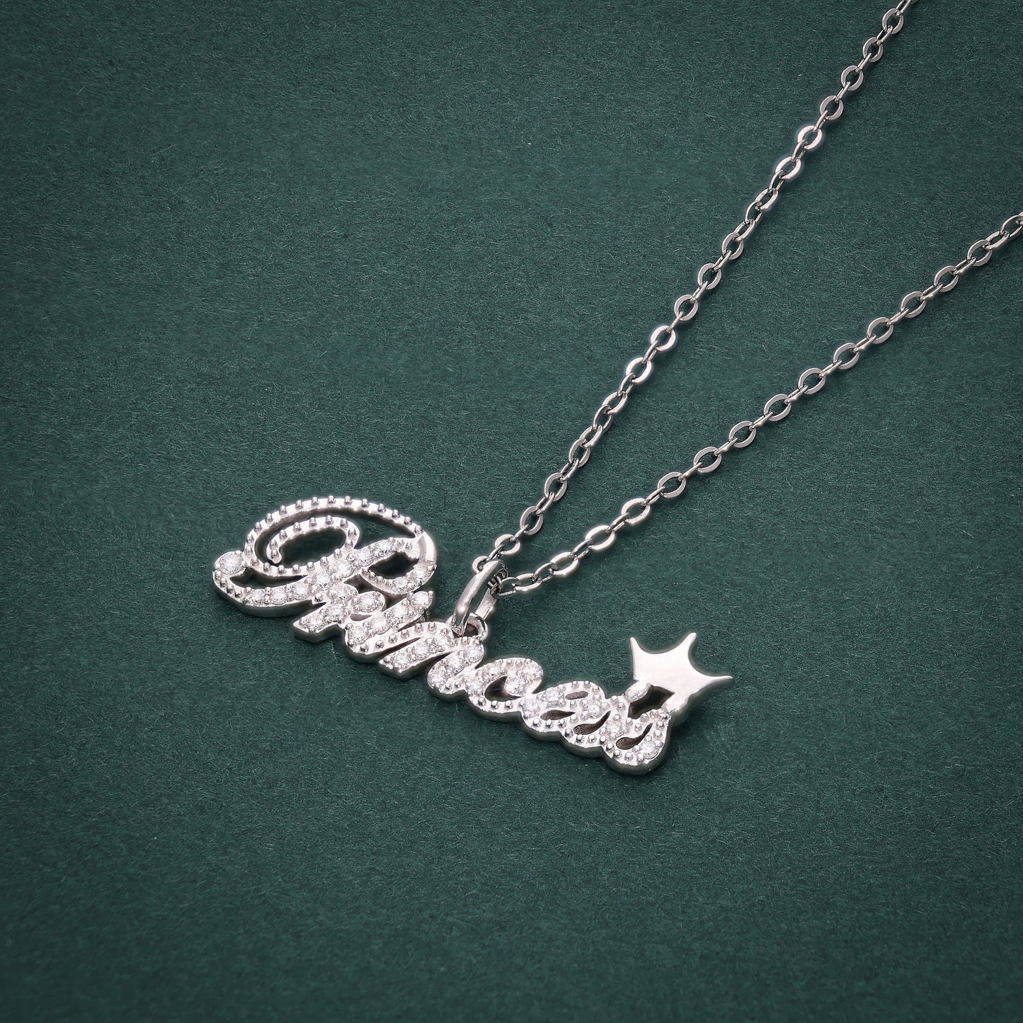A silver pendant with the word 'Princess' written in a cursive script, adorned with diamonds, attached to a silver chain against a green background.