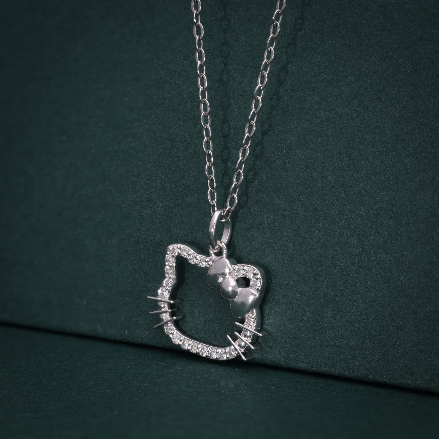 Silver cat face–shaped pendant with lab-grown diamonds, attached to a silver chain and displayed against a dark green background.