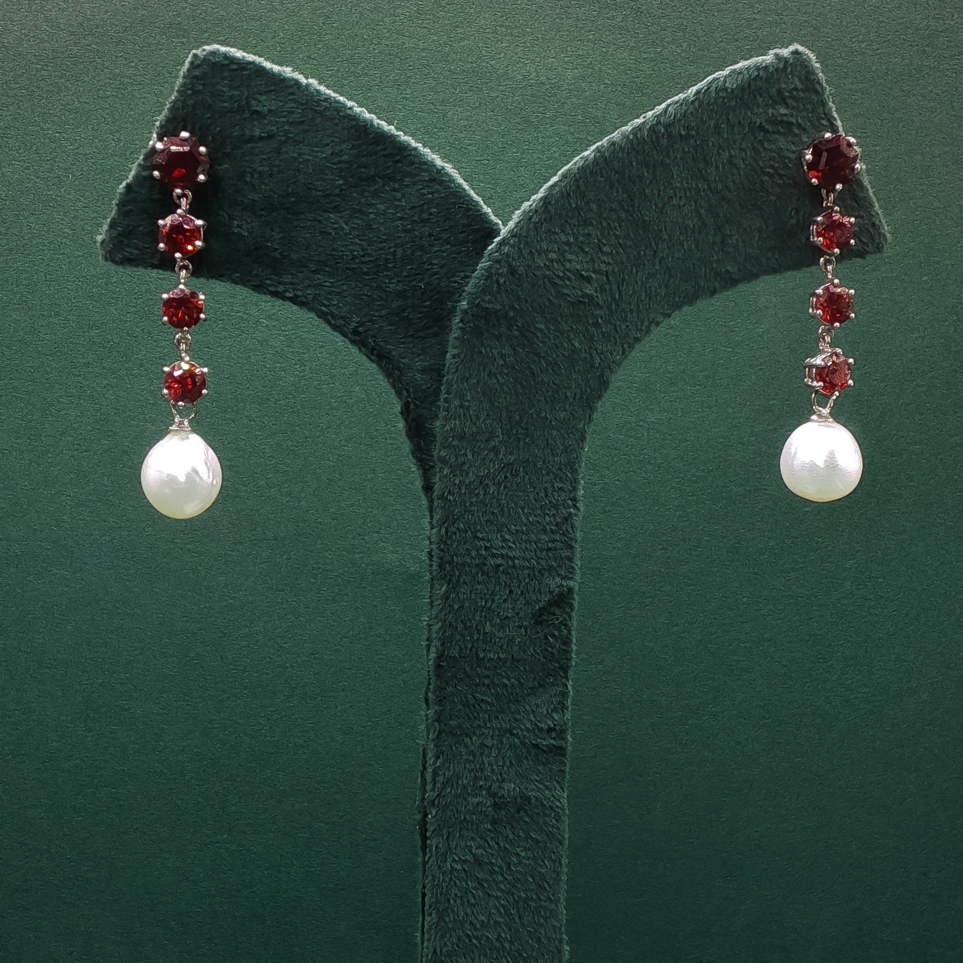 Pair of earrings with pearls and red gemstones on a green stand.
