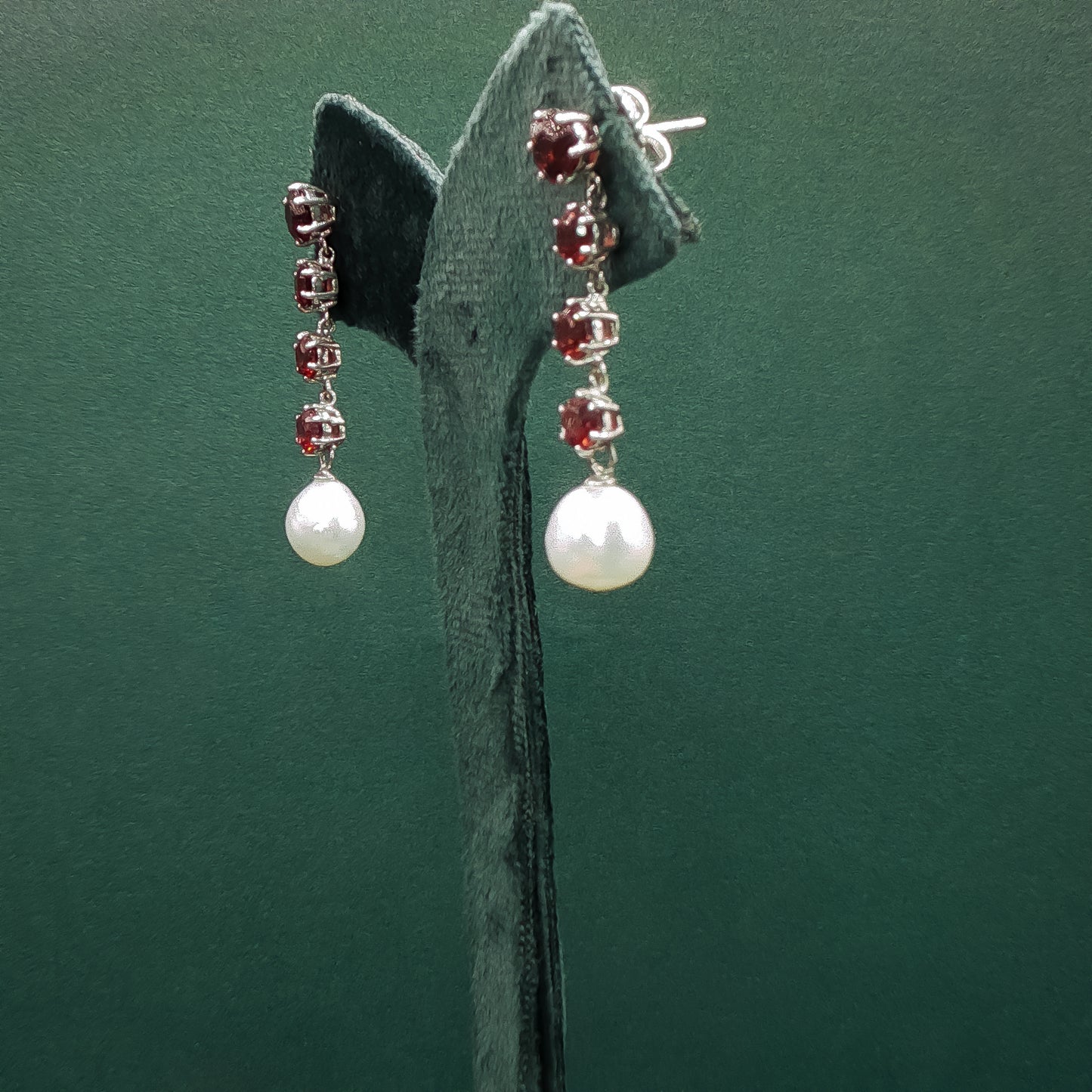 Pair of earrings with pearls and red gemstones on a green stand.