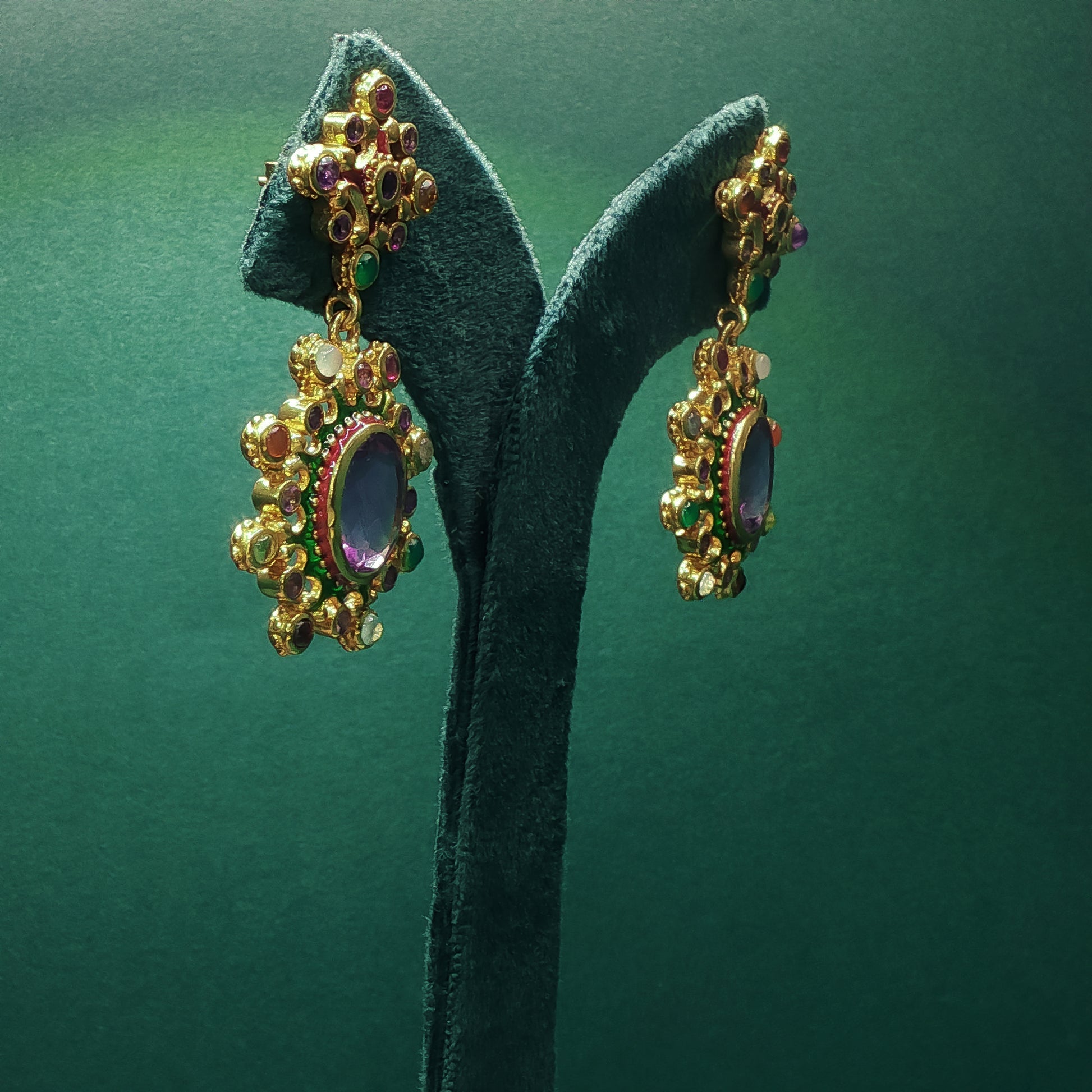 Gold earrings with gemstones on a green stand against a green background

