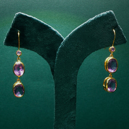 Gold earrings with purple gemstones on a green stand against a green background

