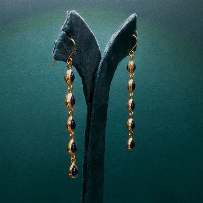 Gold and blue drop earrings on a dark green stand against a matching background